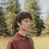 Profile view of a person with curly hair wearing a brown shirt, surrounded by tall grass and trees in a natural setting.