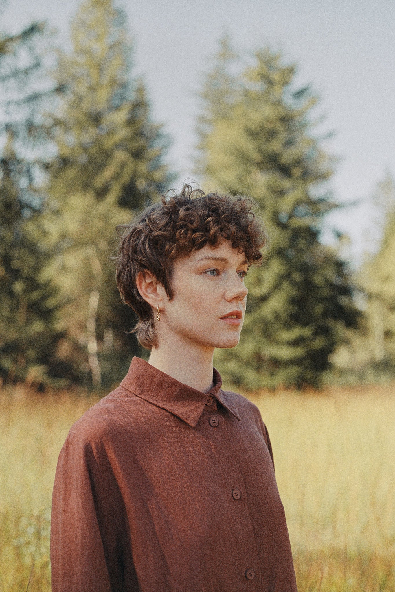 Profile view of a person with curly hair wearing a brown shirt, surrounded by tall grass and trees in a natural setting.