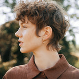 Profile view of a person with curly hair wearing earrings and a brown shirt in a natural outdoor setting.