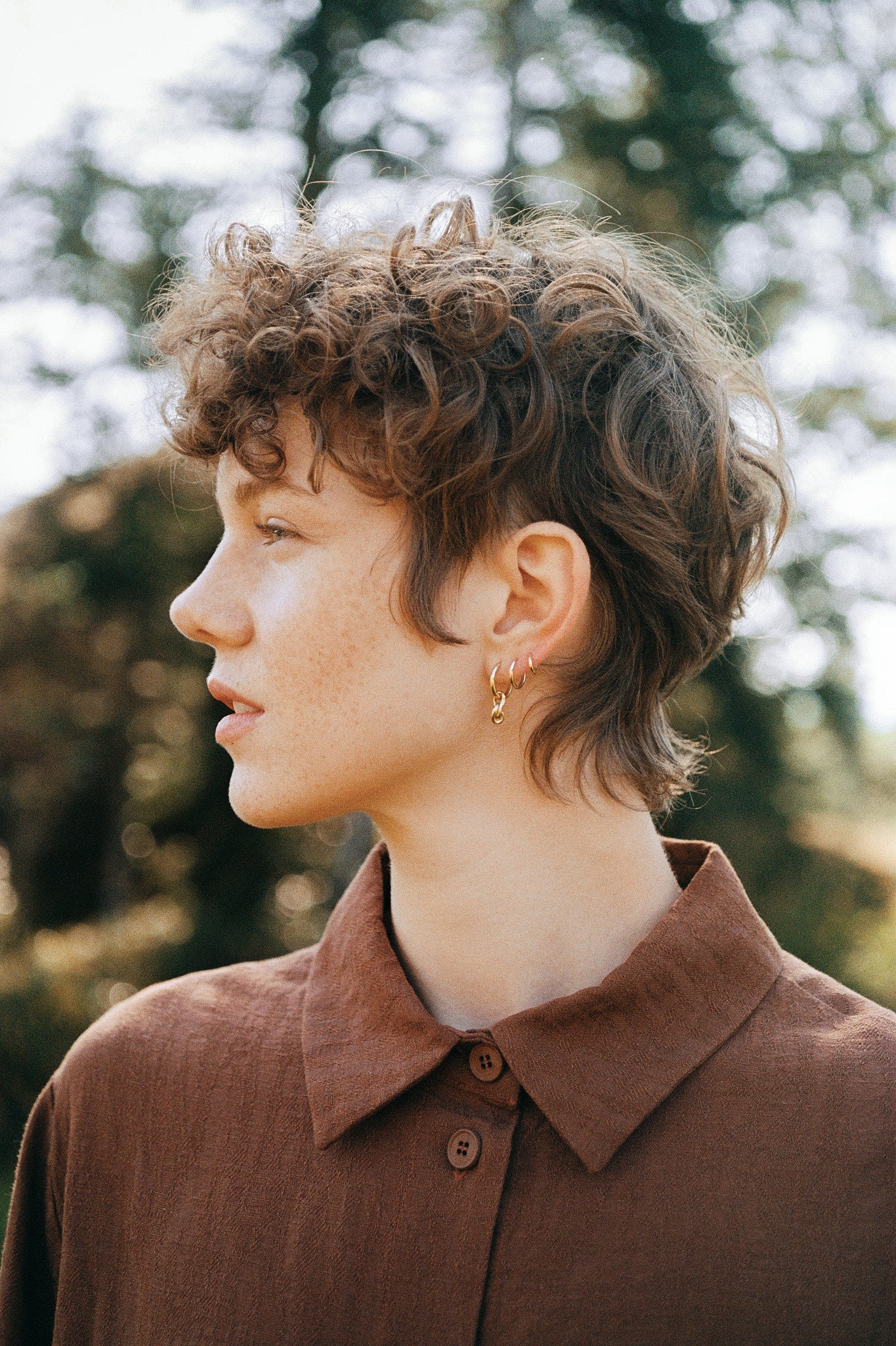 Profile view of a person with curly hair wearing earrings and a brown shirt in a natural outdoor setting.