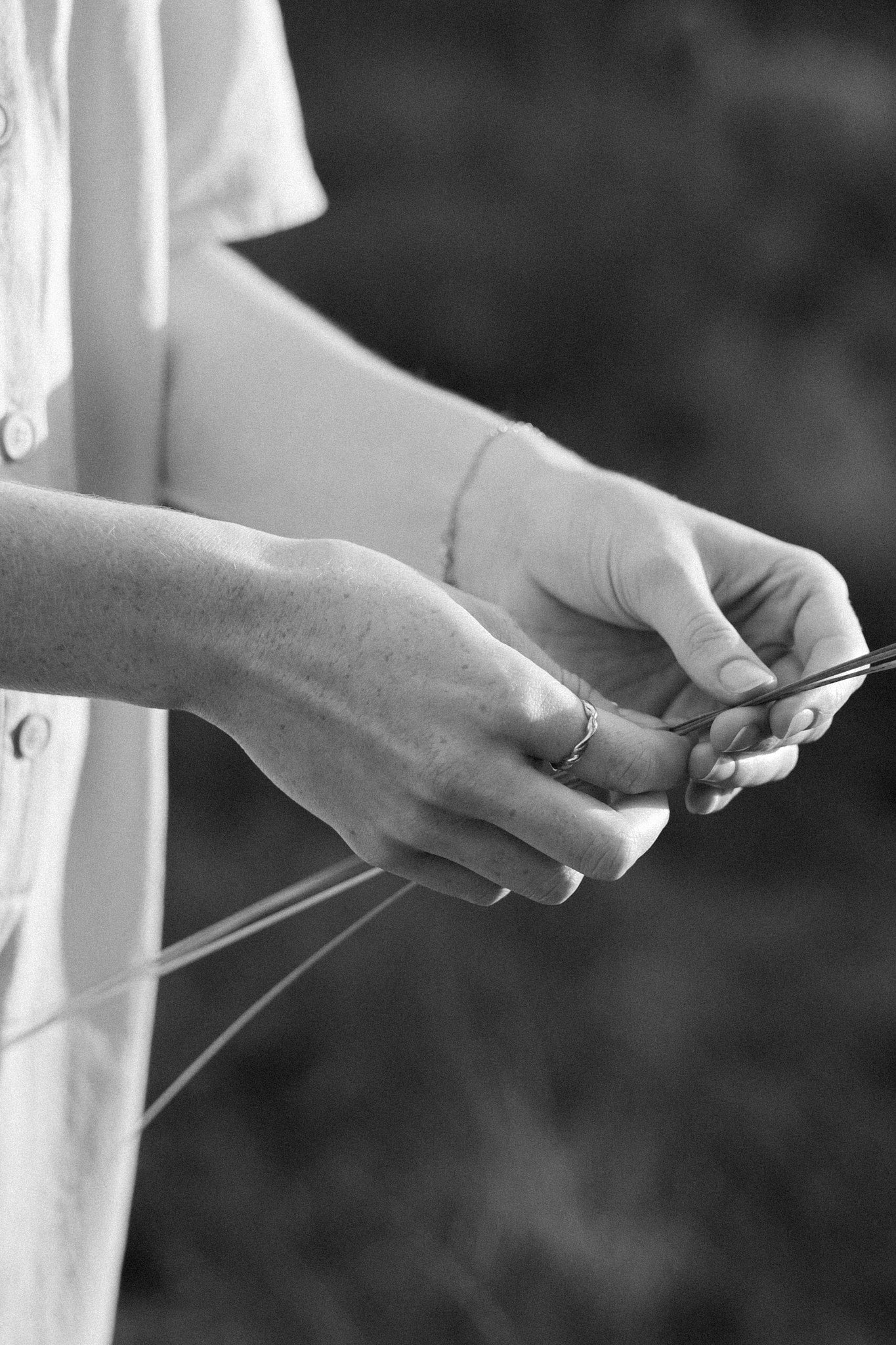Black and white image of hands delicately working with thin strands of material outdoors.