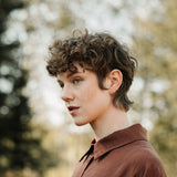 Profile of a person with curly hair wearing a brown shirt, surrounded by blurred greenery.