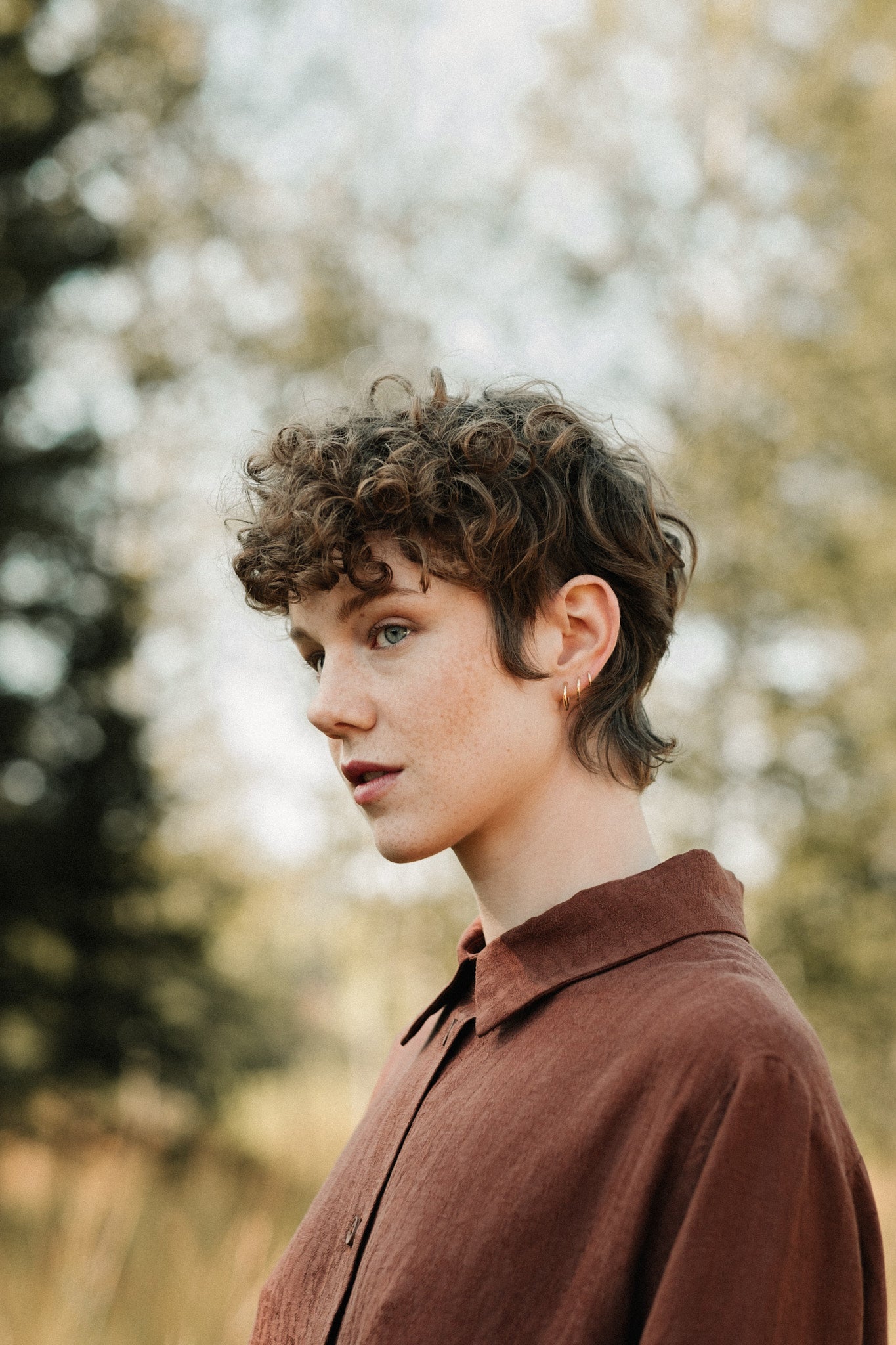 Profile of a person with curly hair wearing a brown shirt, surrounded by blurred greenery.