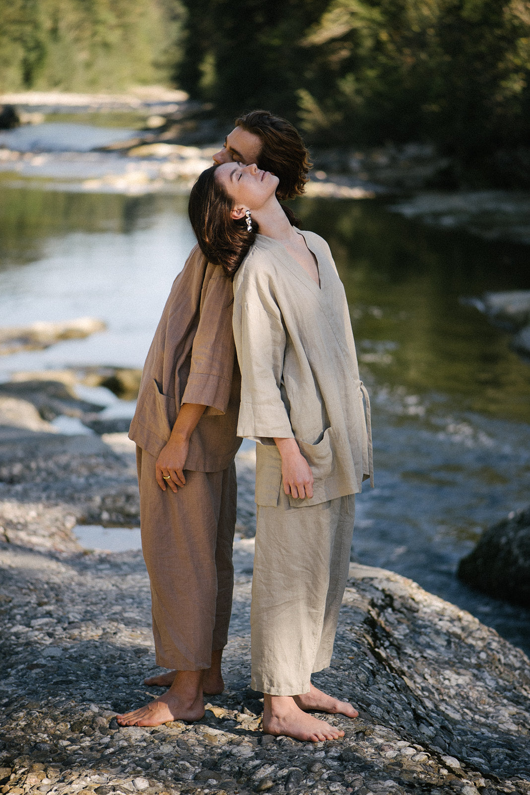 Two models in natural fabrics pose back-to-back by a river, showcasing sustainable fashion styles.