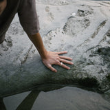 Hand resting on a rock near water, showcasing the DUNA ring made of recycled gold and silver. Ethical jewelry in nature.