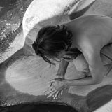 Black and white photo of a person kneeling by water on a rocky surface, hands gently touching the water.