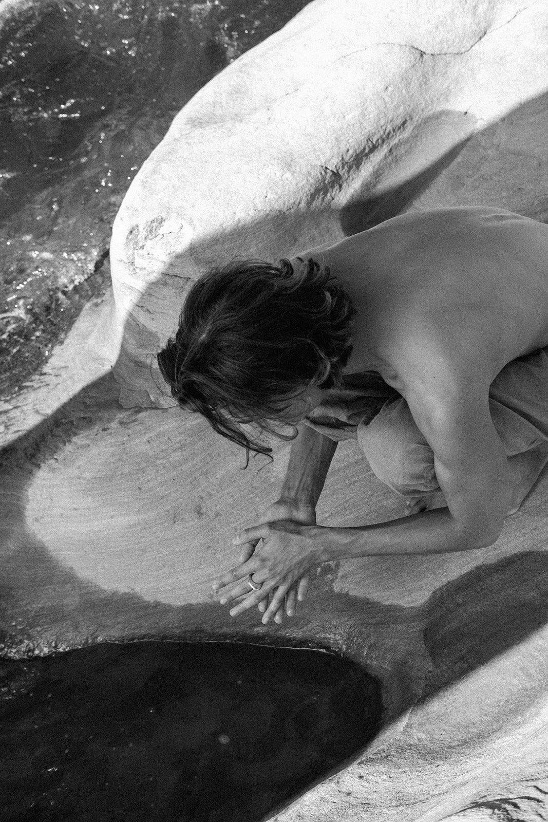 Black and white photo of a person kneeling by water on a rocky surface, hands gently touching the water.