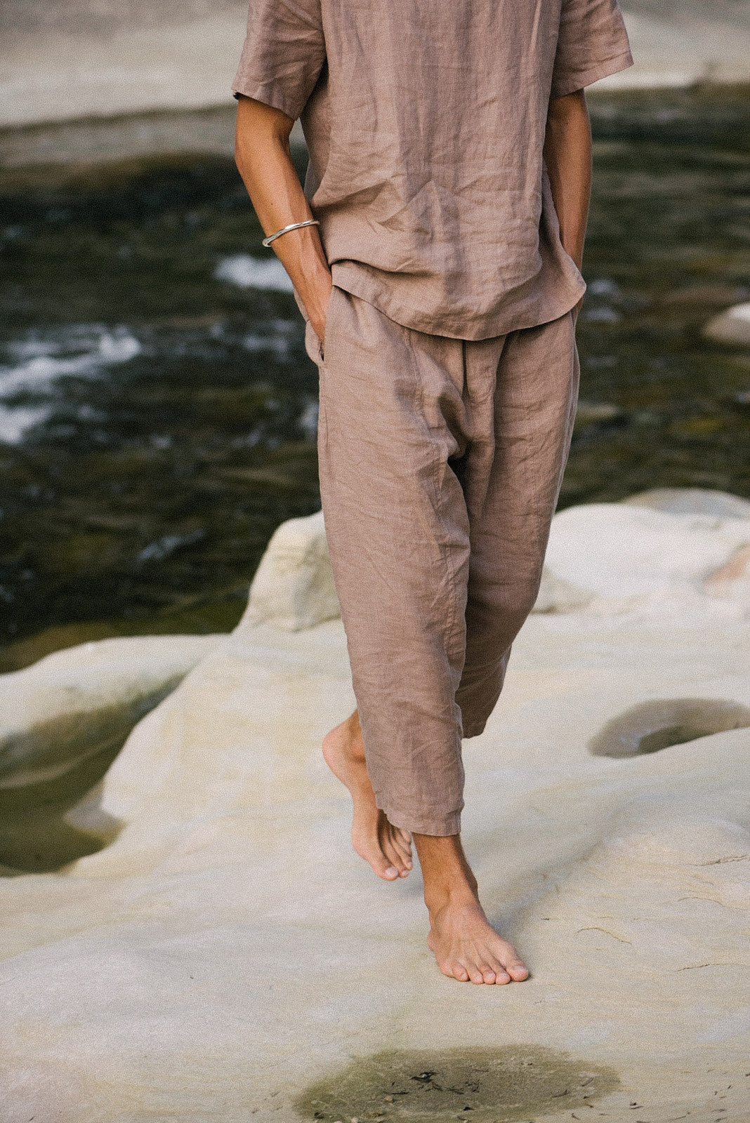 Model wearing a sustainable, handmade linen outfit standing barefoot on a rock by a river.