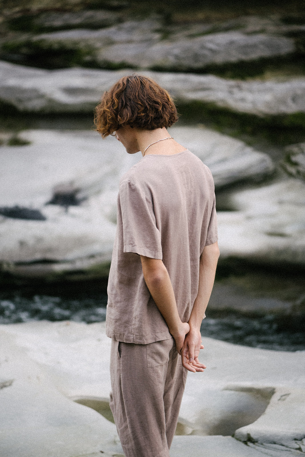 Model wearing sustainable beige outfit standing near water, showcasing an ethical lifestyle in nature.