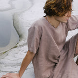 A man with curly hair sits on a rocky surface, wearing a minimalist taupe outfit, reflecting a serene, natural environment.