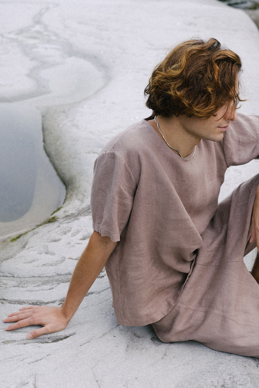 A man with curly hair sits on a rocky surface, wearing a minimalist taupe outfit, reflecting a serene, natural environment.