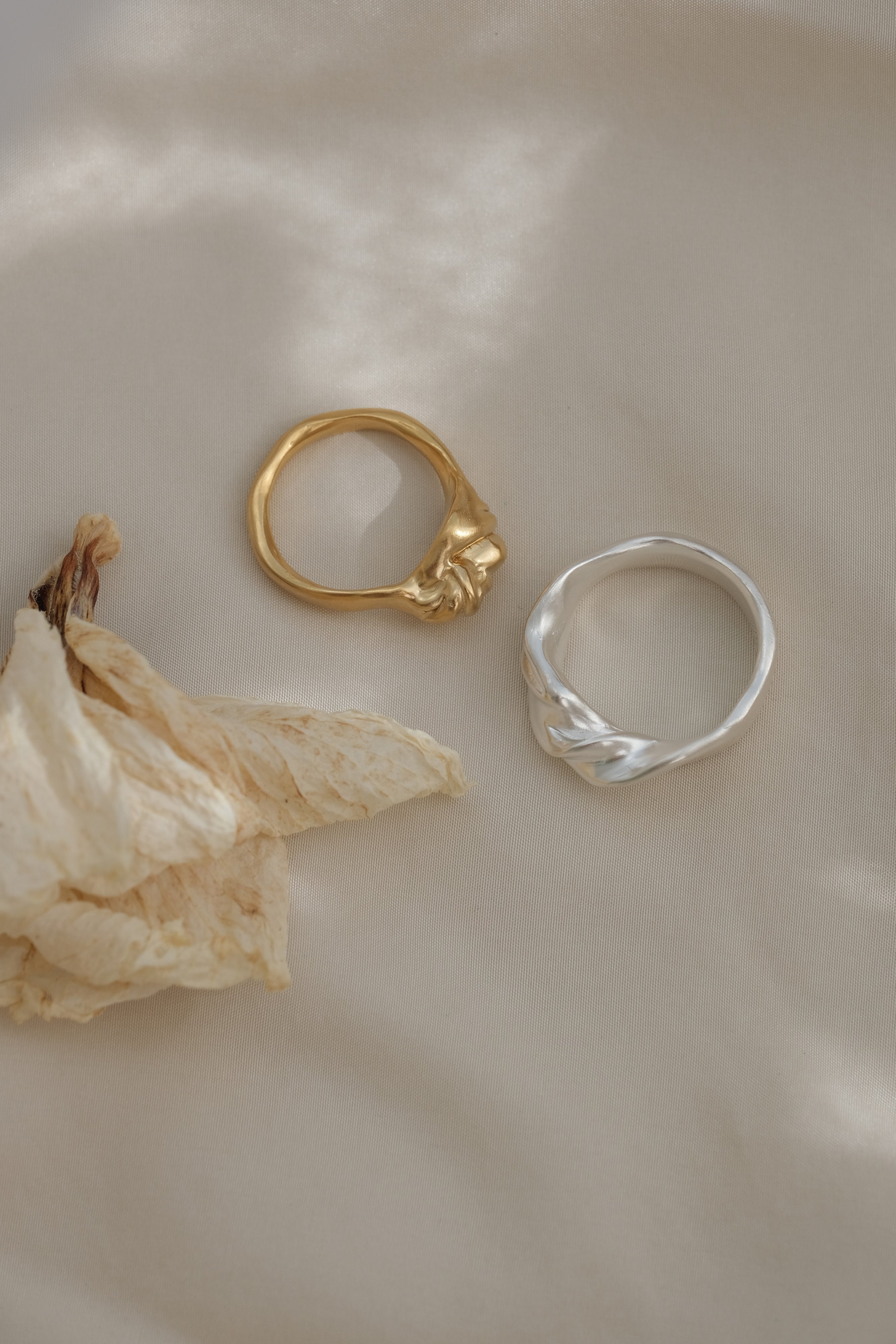 Two handmade unique rings: a gold-plated and a silver ring, against a textured background with dried petals.