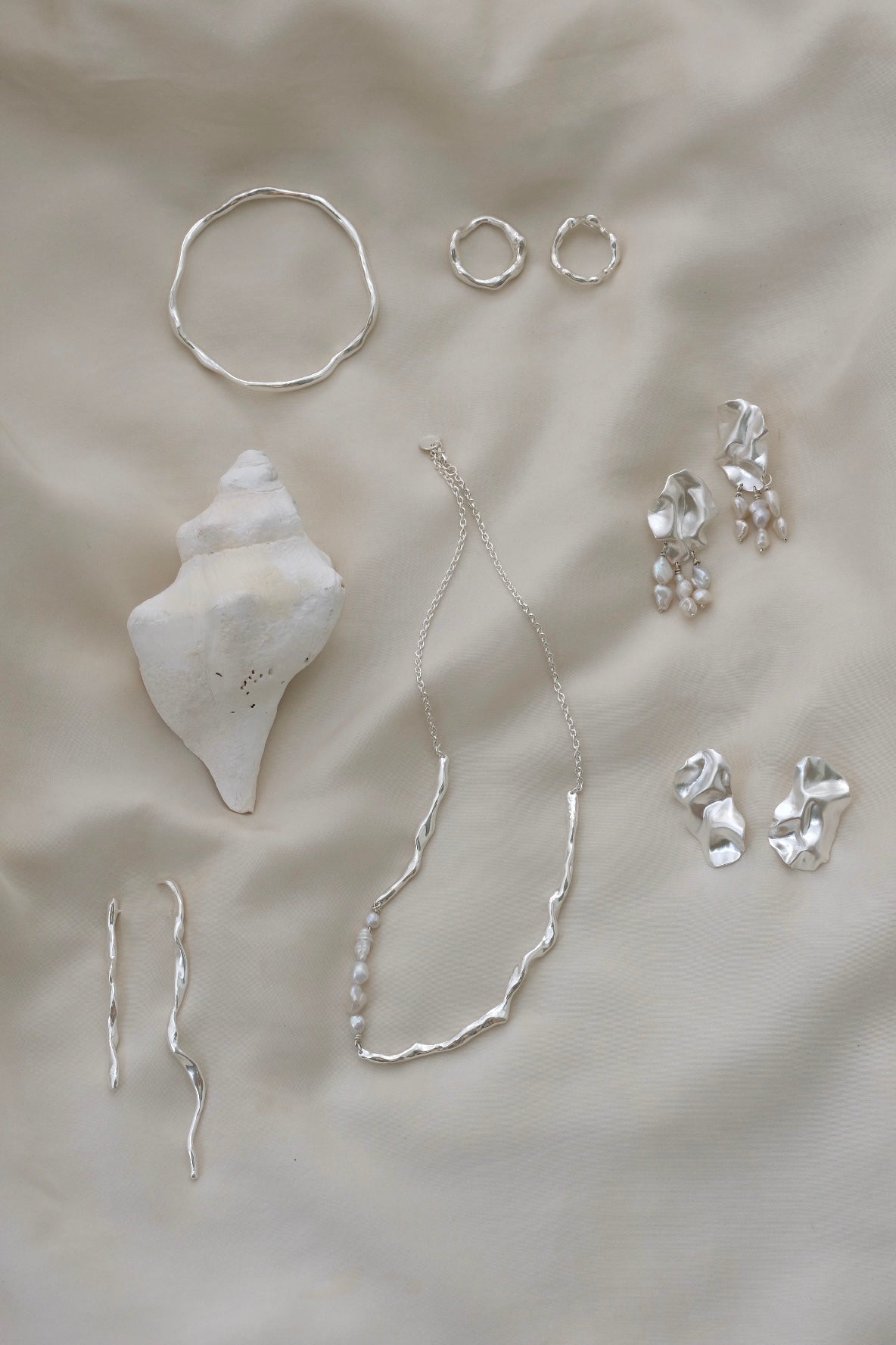 Handmade ethical jewelry collection featuring silver rings, earrings, necklace, and a seashell on a soft surface.