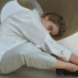 Woman in a white shirt and pants sitting beneath a sculptural structure, showcasing modern fashion in a minimalist setting.