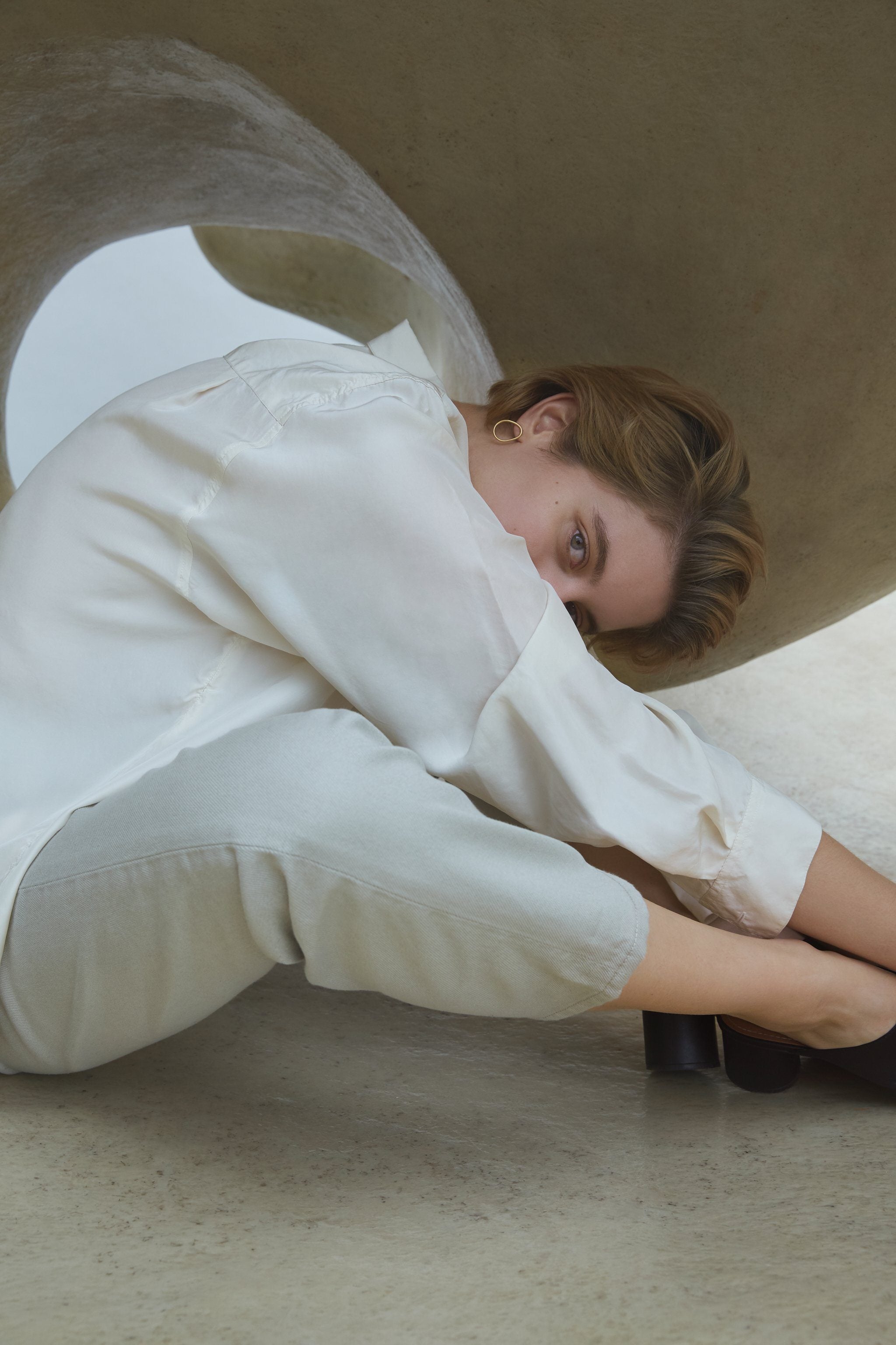 Woman in a white shirt and pants sitting beneath a sculptural structure, showcasing modern fashion in a minimalist setting.