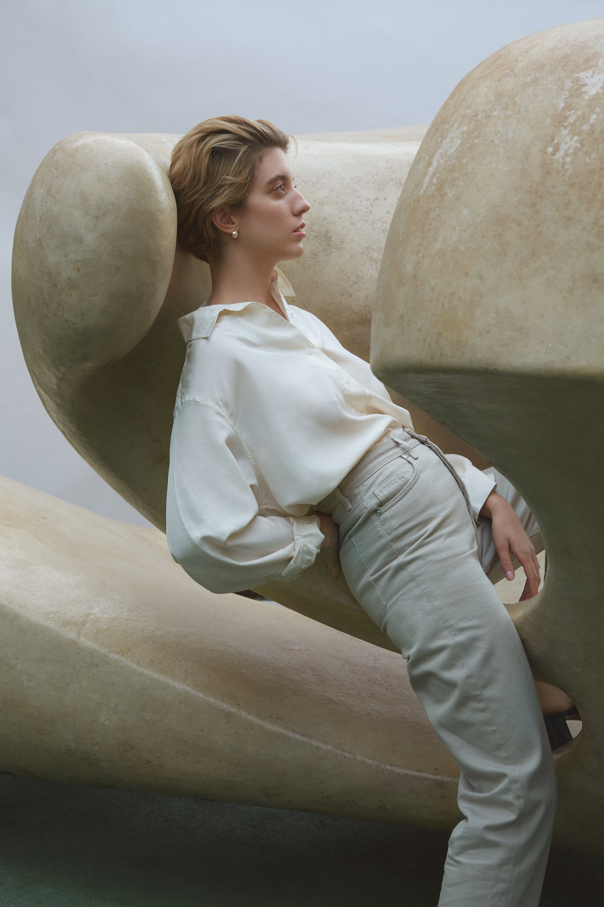 Model wearing a stylish white blouse and beige pants, posed against a sculptural backdrop, showcasing modern fashion design.