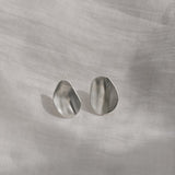 Handcrafted organic earrings in silver on a soft fabric background, showcasing unique shapes.